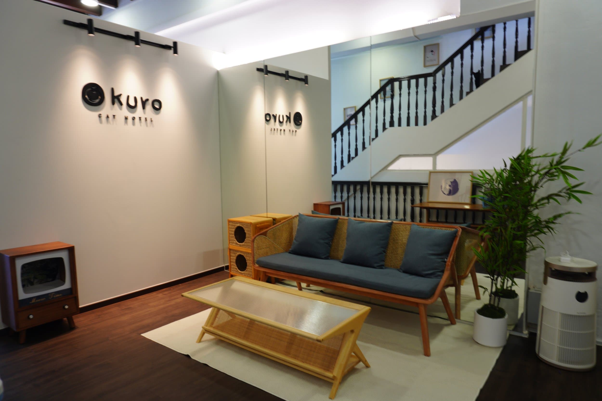 Kuro Cat 猫咪酒店二楼大堂:藤编沙发配蓝色坐垫、矮茶几、印有 Kuro 字样的镜面墙,以及通向三楼的深色木楼梯